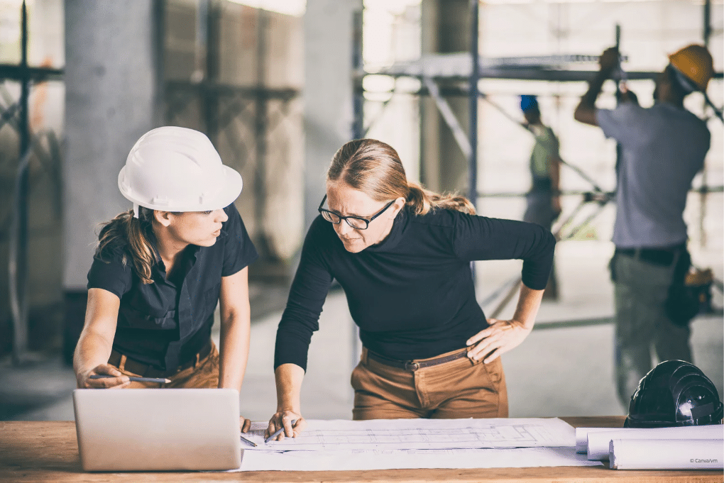 Zwei Frauen stehen an einem Tisch mit Papieren und Laptop und besprechen auf einer Baustelle Pläne.